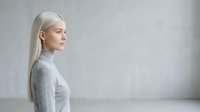 Middle-aged woman with long platinum blonde hair wearing gray turtleneck sweater, profile view against neutral gray background. Suitable for wellness, lifestyle, and professional content.