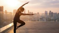 Young woman practicing yoga on urban rooftop terrace at sunrise with city skyline background. Healthy lifestyle and wellness concept for fitness.
