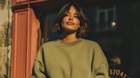Young mixed-race woman in olive green sweatshirt standing outdoors at dusk with warm lighting and urban background for lifestyle and fashion content.