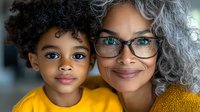 Joyful African American grandmother with gray hair and glasses embracing young granddaughter with curly hair, both smiling warmly at camera. Family bonding moment.