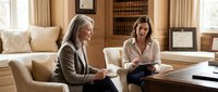 Professional businesswomen discussing strategy in elegant office setting with books and natural light creating productive meeting atmosphere.