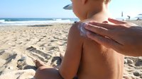 Young boy with tan skin receiving sunscreen application on shoulder at sandy beach with ocean and clear sky in background, suitable for sun protection, family wellness, and summer safety content.