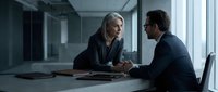 Middle-aged woman with gray hair and man in business suits discussing contract at modern office table with natural light from windows.