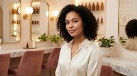 Young African American woman with curly hair smiling in modern beauty salon with pink chairs and warm lighting for wellness and self-care concepts.