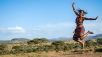 Young woman of African descent wearing traditional patterned clothing, jumping joyfully in savanna landscape with mountains and clear sky, expressing happiness and freedom outdoors.