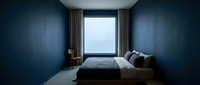 Minimalist bedroom with deep blue walls, large window with gray curtains, low platform bed with dark bedding, wooden chair, and neutral flooring for modern interior design projects.