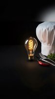 Vintage Edison bulb glowing beside white chef hat with kitchen utensils on dark wooden surface creating warm culinary atmosphere for restaurant branding.