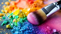 Colorful crushed eyeshadow powder and professional makeup brush on pink background. Scattered rainbow pigments create artistic cosmetic composition for beauty industry.