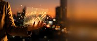 Businessman holding glowing digital tablet displaying financial charts and graphs at sunset with city skyline, representing data analysis, investment growth, and digital technology in business.