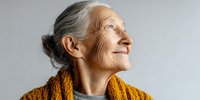Profile of senior Caucasian woman with gray hair in bun, wearing orange knit cardigan, looking up with joyful expression against light gray background.