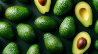 Fresh ripe green avocados and halved avocados with pits on dark background, flat lay composition showing texture and natural pattern of whole fruits.