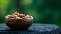 Fresh raw almonds in wooden bowl on dark stone surface against blurred green natural background, healthy organic snack food photography.
