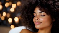 Young African American woman with natural curly hair and glowing skin against blurred golden bokeh lights background, eyes closed with serene peaceful expression.