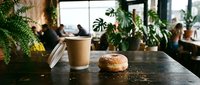 Coffee cup with pastry on dark table in modern cafe with lush green plants and blurred customers in background, suitable for hospitality, food and beverage, and lifestyle marketing.