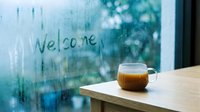 Steaming coffee mug on wooden table near foggy window with Welcome written in condensation, cozy rainy day atmosphere.