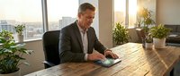 Middle-aged Caucasian male professional in business casual attire working at wooden desk with computer mouse in modern office with city skyline view and potted plants.