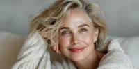 Mature blonde woman in cozy sweater smiling warmly with hands in hair, expressing confidence and natural beauty for lifestyle and wellness content.