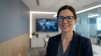 Professional businesswoman with glasses smiling in modern office environment with conference room and technology displays in background for corporate presentations.