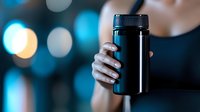 Hand holding black reusable water bottle or thermos against blurred bokeh background in blue tones, sustainable lifestyle and eco-friendly concept.