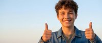 Happy teenage boy with curly hair giving thumbs up gesture wearing denim jacket against clear sky background expressing positive emotions and success.