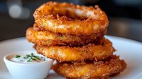 Crispy golden onion rings stacked on white plate with creamy dipping sauce, garnished with fresh herbs. Close-up shot showing crunchy breaded texture.