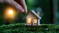 Small wooden house model with glowing windows placed on green moss at sunset, creating magical miniature scene with bokeh light effect in background.