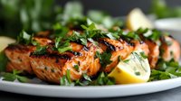 Grilled salmon fillets garnished with fresh herbs and lemon on white plate, close up view showing moist, flaky texture and caramelized surface. For food blogs and menus.