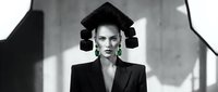 Young Caucasian woman in avant-garde black suit with sculptural hair and green earrings, monochrome fashion editorial portrait.