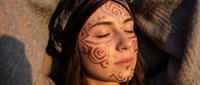 Young woman with traditional tribal face paint patterns relaxing on sandy beach in warm sunlight with closed eyes and peaceful expression.