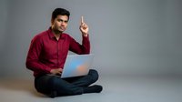 Young Indian businessman in burgundy shirt sitting cross-legged with laptop pointing up having eureka moment on gray background for business concepts.