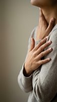 Woman touching neck and chest area with hands in beige sweater showing throat pain or respiratory discomfort for health and wellness concepts.