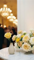 Elegant cream and yellow rose bouquet in ceramic vase on marble table with warm chandelier bokeh lights creating luxurious restaurant ambiance.
