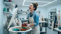 Young female potter laughing joyfully while working on pottery wheel in bright ceramic studio workshop. Creative artisan crafting clay bowls in modern art space.