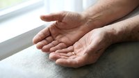 Senior man cupped hands gesture showing open palms in prayer or receiving position on soft fabric surface near window with natural lighting.