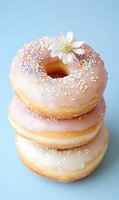 Stack of pink glazed donuts with colorful sprinkles and white flower decoration on pastel blue background for bakery and dessert concepts.