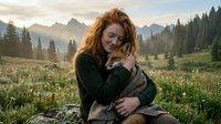 Young redhead woman cuddling small dog in mountain meadow during golden hour sunset with alpine peaks and forest background for outdoor lifestyle content.