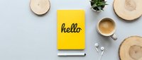 Yellow notebook with hello lettering, coffee cup, earphones and plant on light gray background, flat lay workspace top view.
