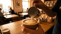 Barista pouring steamed milk creating latte art in coffee cup at cozy cafe counter. Professional coffee making process in warm restaurant atmosphere.