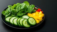 Fresh green spinach leaves, sliced cucumber rounds, and yellow bell pepper arranged on black plate against dark background. Healthy vegetarian meal.