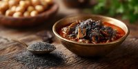 Spicy curry dish with tender meat in copper bowl, black sesame seeds on wooden table. Traditional Asian cuisine with rich sauce and aromatic spices.