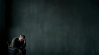 Stressed middle-aged man with dark hair and beard, wearing business suit, sitting in contemplative pose against dark textured wall. Suitable for mental health, depression, anxiety, and corporate stress concepts.