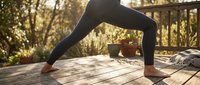 Woman in downward dog yoga pose on wooden deck surrounded by natural sunlight and forest scenery, practicing fitness and wellness outdoors.