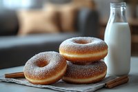Fresh sugar-dusted donuts stacked on linen napkin with glass bottle of milk and cinnamon sticks, creating cozy breakfast atmosphere on wooden surface.