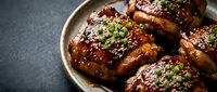 Glazed chicken thighs with sesame seeds and fresh herbs on ceramic plate. Caramelized poultry dish with Asian-inspired sauce for restaurant menu photography.
