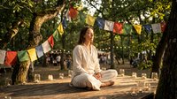 Young woman in white clothing meditating peacefully outdoors under colorful prayer flags strung between trees in serene garden setting with warm sunlight.