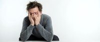 Stressed young Caucasian man with messy hair covering face with hands while sitting at desk, expressing frustration, burnout or mental health struggles.