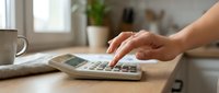 Woman calculating finances on calculator at home office desk with coffee mug and notebook for budgeting planning and accounting tasks.