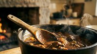 Wooden spoon stirring hot stew in cast iron pot with steam rising, fireplace glowing in background, cooking and food preparation.