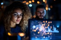 Young mixed-race woman with curly hair and glasses analyzing financial data on computer screen with male colleague in modern office at night.