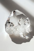 Clear quartz crystal cluster with sharp geometric formations catching light and casting shadows on white surface, raw mineral specimen in natural form.